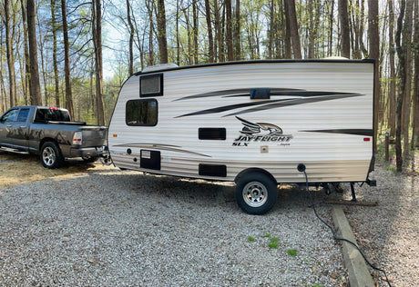 RV rental setup: A Jayco travel trailer hitched to a pickup truck on a gravel campsite amidst trees, perfect for camping.