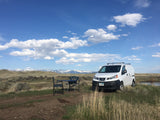 White Nissan van with chairs/table set up, facing snow-capped mountains under blue sky. Ideal RV rental for scenic adventures.