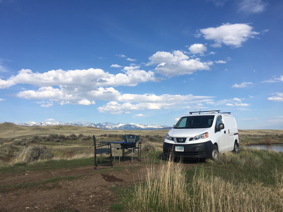 White Nissan van with chairs/table set up, facing snow-capped mountains under blue sky. Ideal RV rental for scenic adventures.