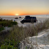 Black camper van parked on a sandy beach at sunset, showcasing the freedom of an RV rental for coastal adventures.