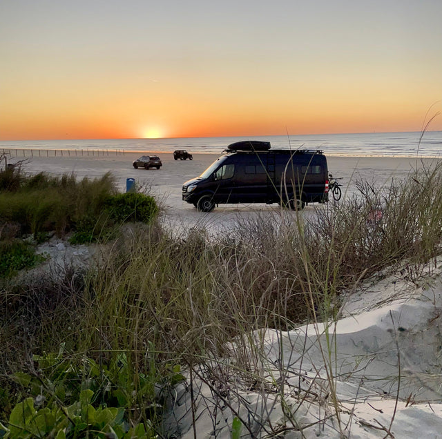 Black camper van parked on a sandy beach at sunset, showcasing the freedom of an RV rental for coastal adventures.