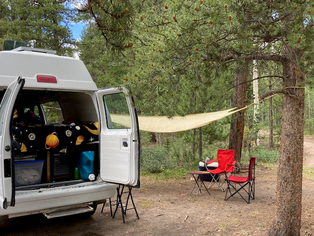 RV rental camping setup: A white camper van, bed with space sheets, shade sail, and chairs in a forest campsite.