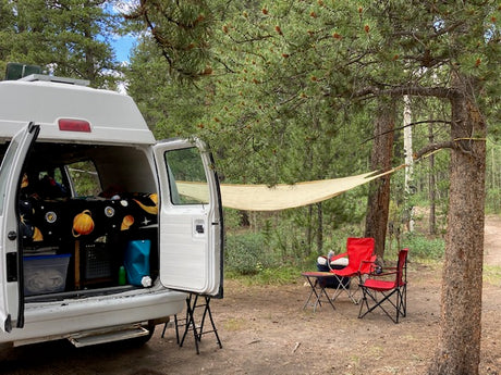 RV rental camping setup: A white camper van, bed with space sheets, shade sail, and chairs in a forest campsite.