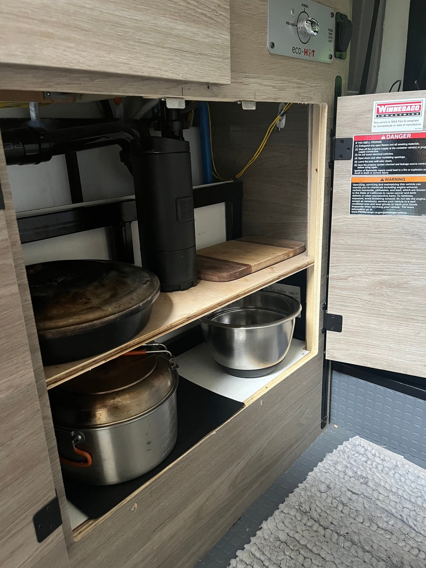 RV rental interior: organized under-counter storage with cookware like pots, pans, and a cutting board. Winnebago labels. — photo 12