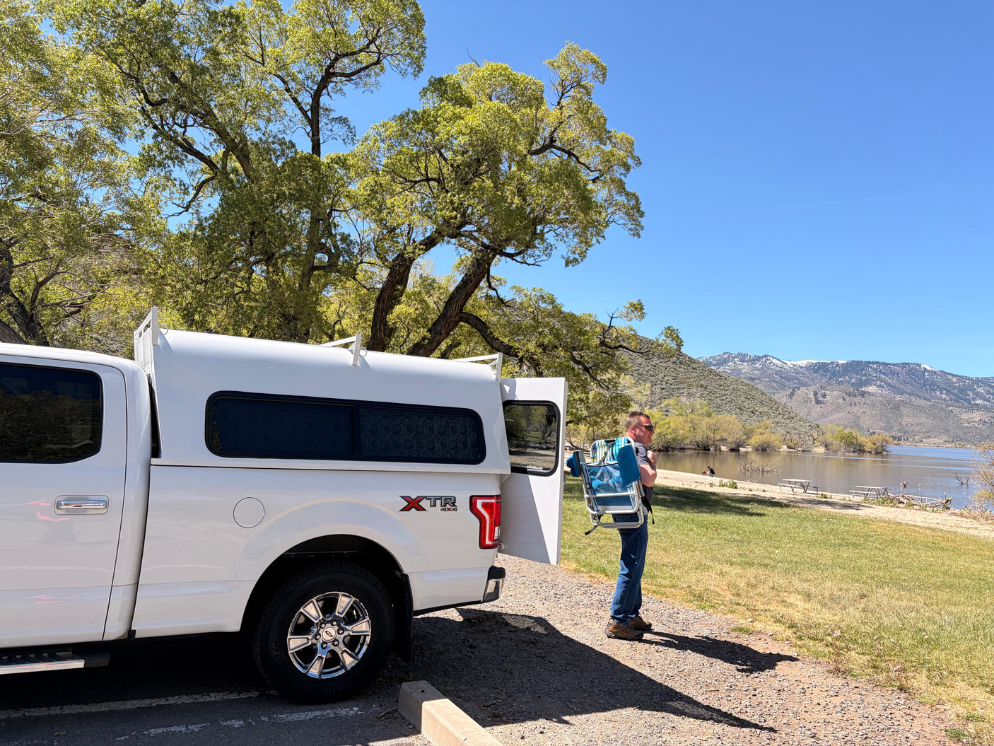 Man with beach chair by a lake & mountains, next to a white pickup truck with a camper shell, perfect for an RV rental adventure.