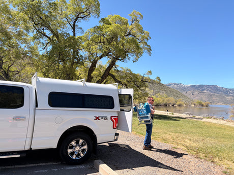 Man with beach chair by a lake & mountains, next to a white pickup truck with a camper shell, perfect for an RV rental adventure.