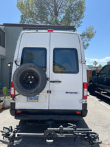 Rear view of a white Mercedes Sprinter van, a common RV rental. It has a spare tire, bike rack, and roof-mounted solar panels.