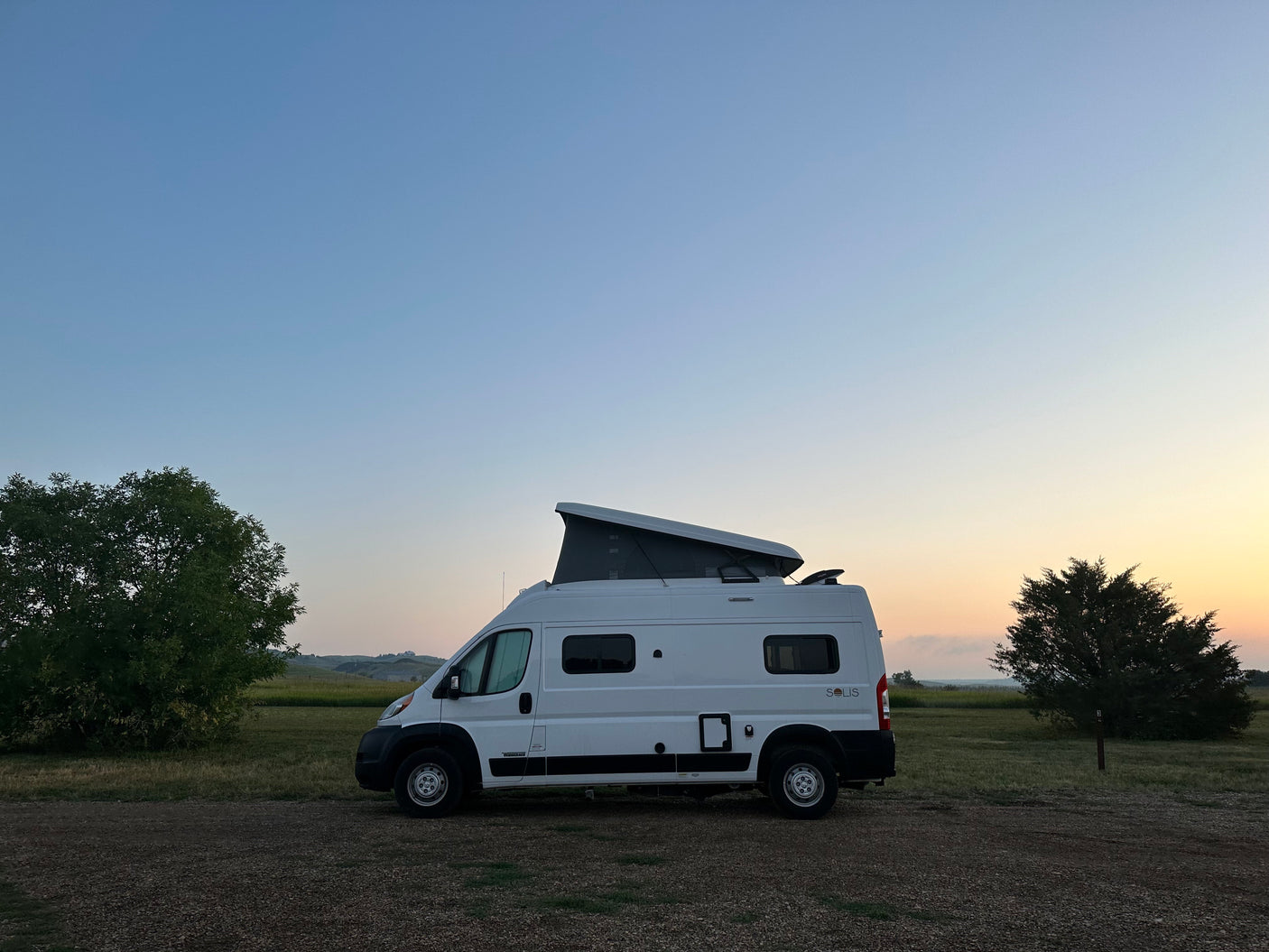 White Winnebago Solis RV rental camper van with pop-top open, parked in a scenic field during golden hour.