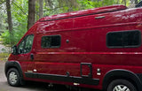 A beautiful red camper van, ready for adventure! This rv rental is parked in a lush, green wooded area, perfect for travel.