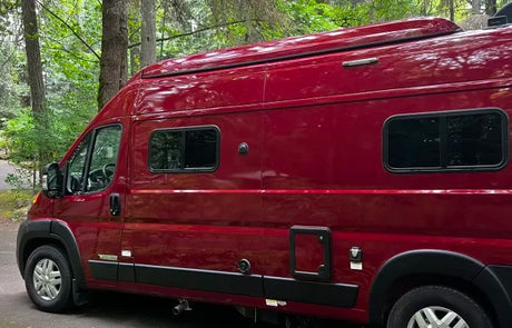 A beautiful red camper van, ready for adventure! This rv rental is parked in a lush, green wooded area, perfect for travel.