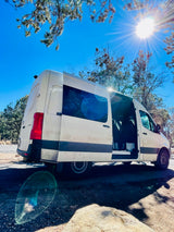 Bright sun illuminates this white Mercedes-Benz Sprinter rv rental, parked with doors open in a natural, tree-lined setting.