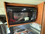RV rental kitchen features a sleek black microwave oven, integrated into wooden cabinetry above a metallic counter. Shows user reflection.
