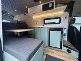 Modern RV rental interior features a mint green kitchenette with a sink, microwave, and fridge, adjacent to a bed and dining area.