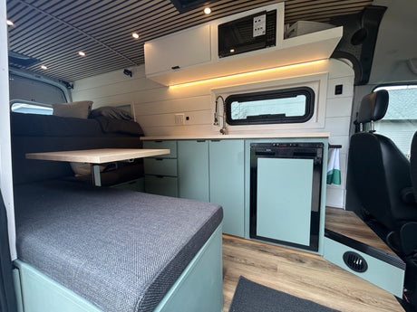 Modern RV rental interior features a mint green kitchenette with a sink, microwave, and fridge, adjacent to a bed and dining area.