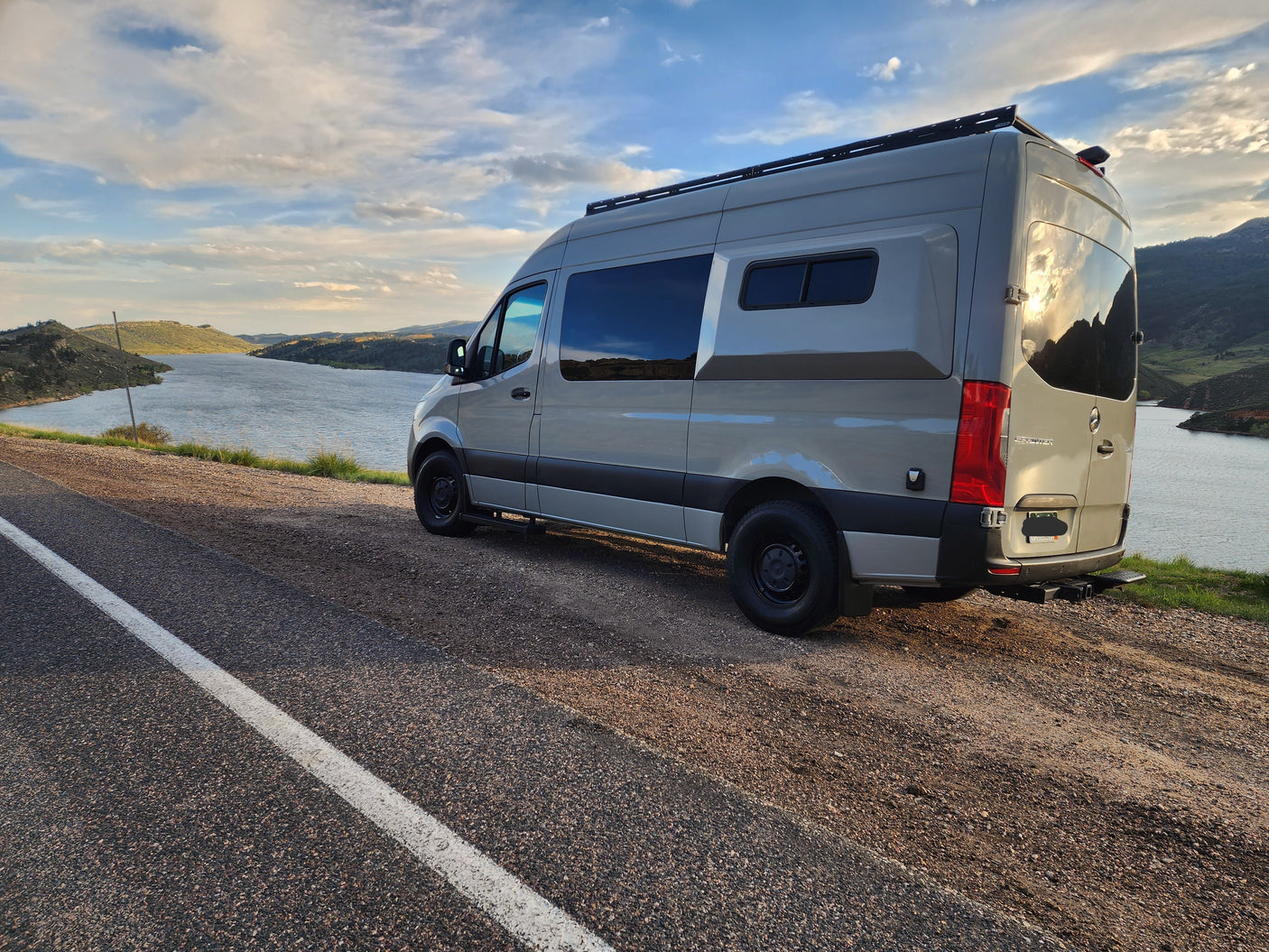 Light gray RV rental camper van parked on a scenic lakeside road, mountains under a cloudy sky. Perfect for a road trip.