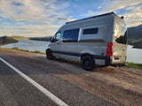 Light gray RV rental camper van parked on a scenic lakeside road, mountains under a cloudy sky. Perfect for a road trip.