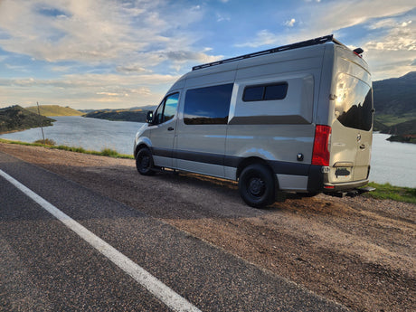 Light gray RV rental camper van parked on a scenic lakeside road, mountains under a cloudy sky. Perfect for a road trip.