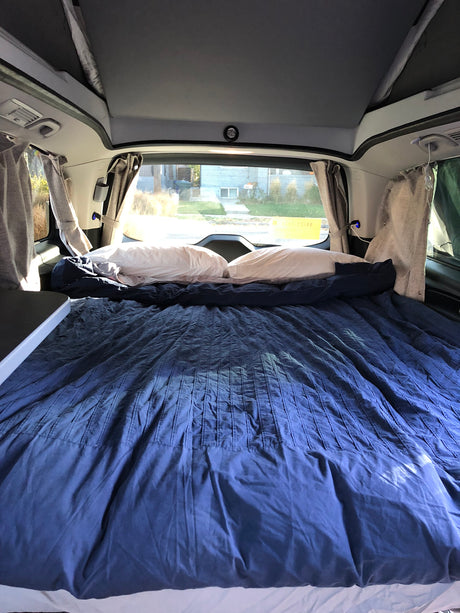 Inside a cozy RV rental: a large bed with blue bedding and pillows, curtains, and a view outside, ideal for campervan adventures.