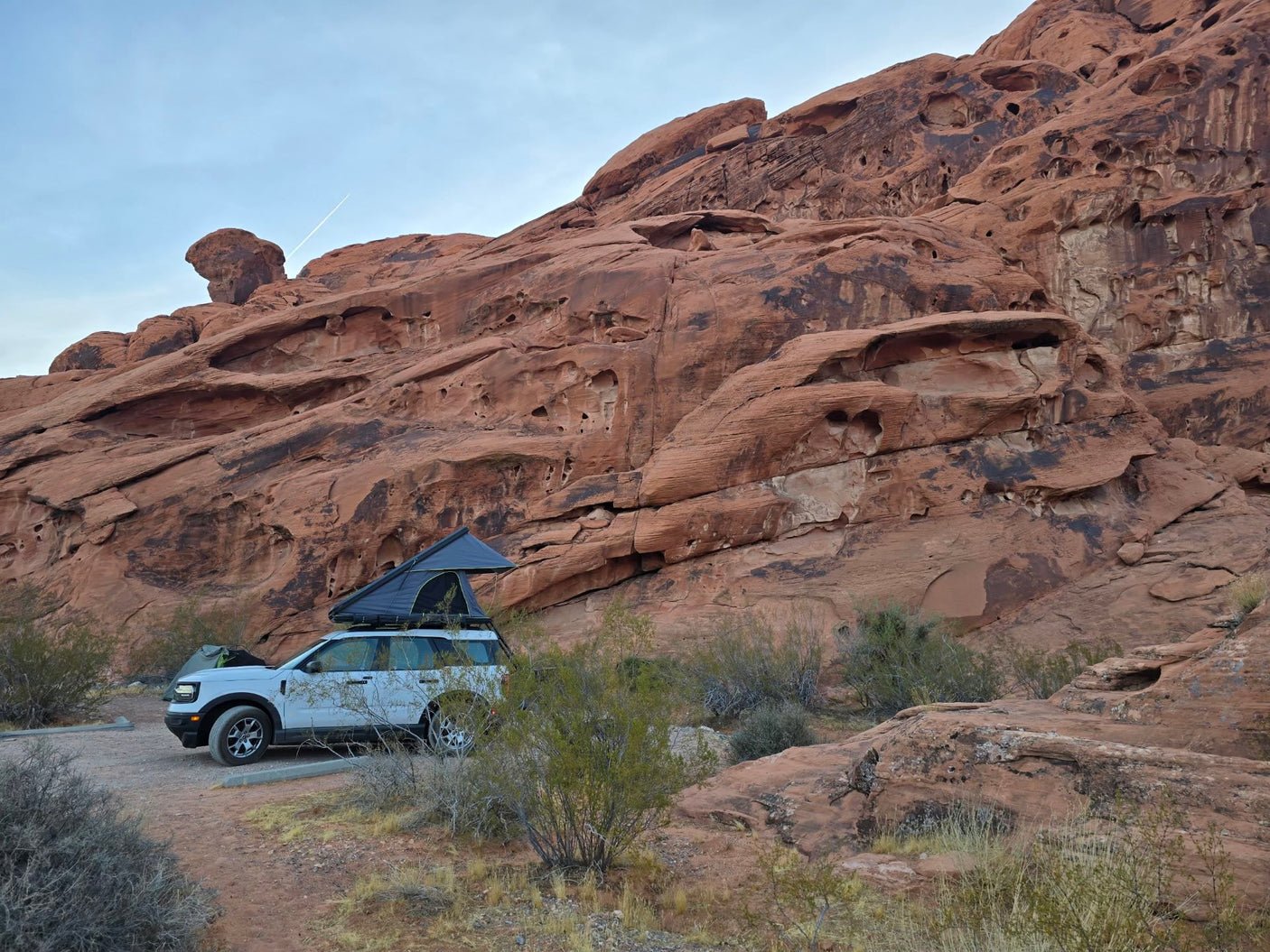An rv rental-style setup: white SUV with rooftop tent parked at red rock cliffs, ready for desert camping.