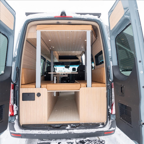 Interior of an RV rental camper van: light wood, tan cushions forming U-shaped seating/bed, table, seen from rear in snow.