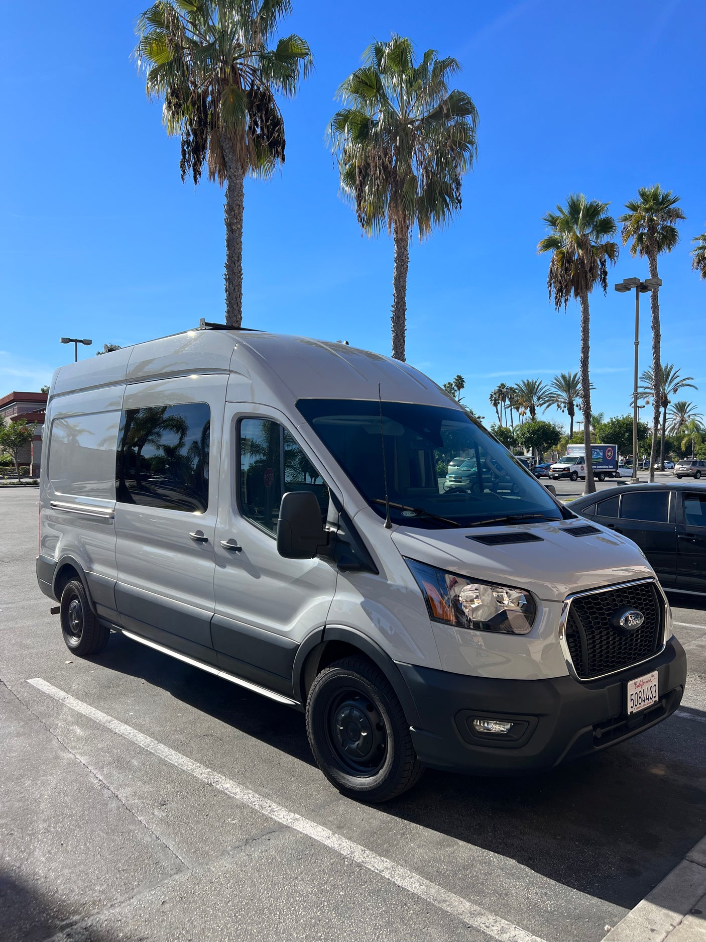 RV rental: Light grey Ford Transit camper van in a sunny parking lot with palm trees.
