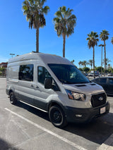 RV rental: Light grey Ford Transit camper van in a sunny parking lot with palm trees.