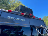 This RV rental's rear features a rooftop AC, "SEQUENCE" lettering, rear lights, and a bike rack against a blue sky.