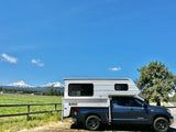 RV rental: A grey truck with a white truck camper parked by a fence, facing snow-capped mountains & green fields.