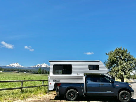 RV rental: A grey truck with a white truck camper parked by a fence, facing snow-capped mountains & green fields.