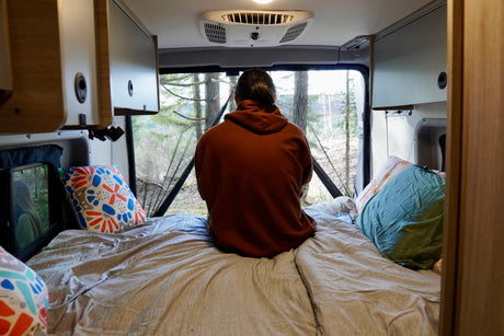 Cozy RV rental bed with a person enjoying the serene forest view. Ideal for nature escape and van life adventure.