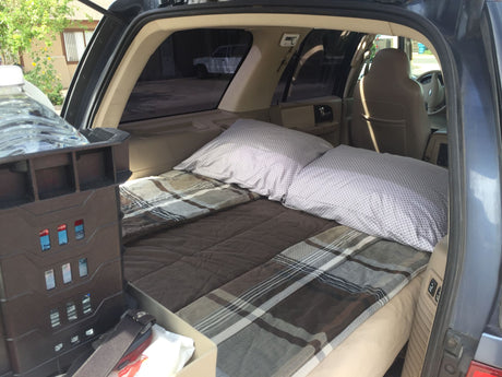 This cozy RV rental car interior features a bed with pillows and plaid blankets in the cargo area, with a storage unit visible.