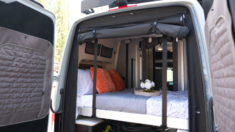 Cozy RV rental bed in a camper van with open rear doors revealing nature. Perfect for van life travel.