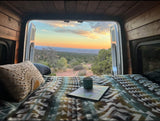 Scenic RV rental adventure: cozy bed setup with a stunning sunset over the landscape, seen from inside the van.