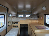 Inside this modern rv rental, a custom camper van features a bed, compact kitchen, and lounge area with light wood panels.