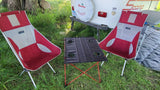 Two red camping chairs and a table are set up on grass, next to a white Scamp RV rental.