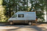 Experience outdoor living with an rv rental! A person relaxes on a rooftop deck of a grey camper van surrounded by trees.