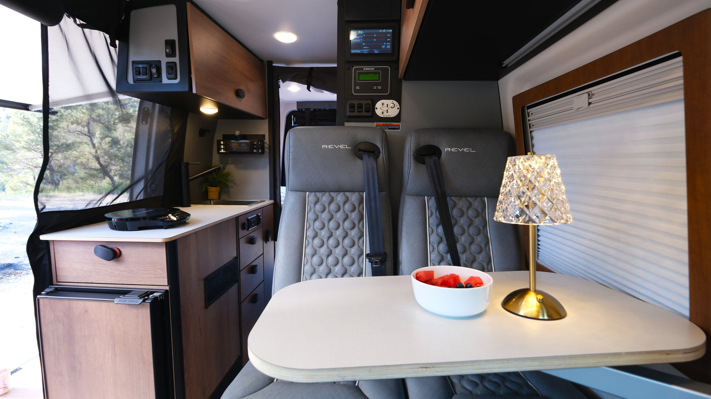 Cozy RV rental interior: dining area with two seats, a table with fruit and lamp, and a compact kitchenette.