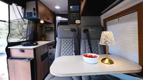 Cozy RV rental interior: dining area with two seats, a table with fruit and lamp, and a compact kitchenette.