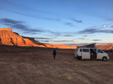 Desert sunset with an RV rental camper van parked and a person enjoying the view of the glowing mountains and moon.