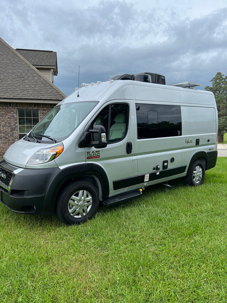 Silver Ram ProMaster camper van parked on green grass, ready as an RV rental. Visible features include a deployed step and roof units.