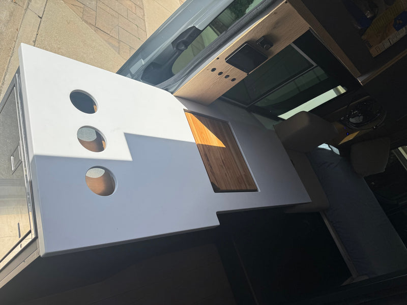 RV rental: Custom camper van interior with a white countertop, round cutouts, wood insert, & overhead controls. — photo 11