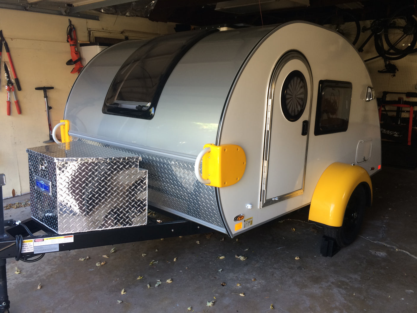 A modern teardrop camper with a shiny front diamond plate storage box and yellow fenders, suitable for an RV rental.