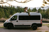 Woman on a camper van roof enjoying mountain views. Explore nature with an RV rental for your next adventure.