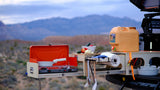 RV rental outdoor kitchen setup: camp stove, water dispenser, fresh veggies, utensils, and paper towels with a mountain view.