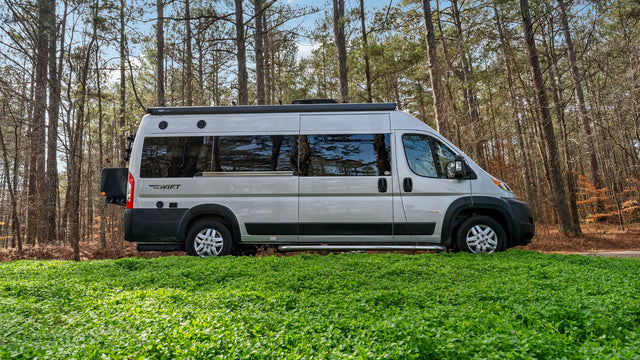 Silver camper van RV rental on vibrant green foliage by tall pine trees, ready for outdoor adventures. Jayco Swift model.