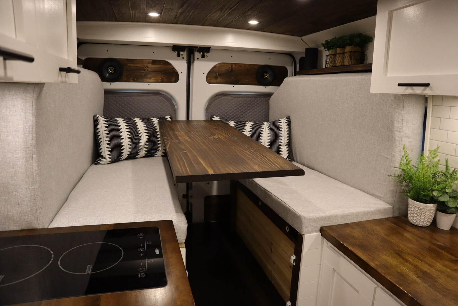 Spacious RV rental camper van interior. Features light grey benches, a wooden table, kitchen counter with cooktop, and storage.