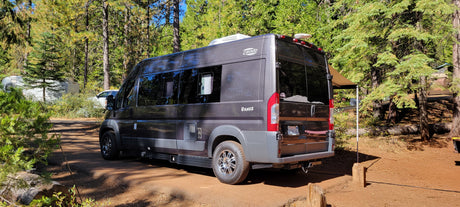 This RV rental camper van is parked in a sunny, wooded campground with tall trees, ideal for outdoor travel.