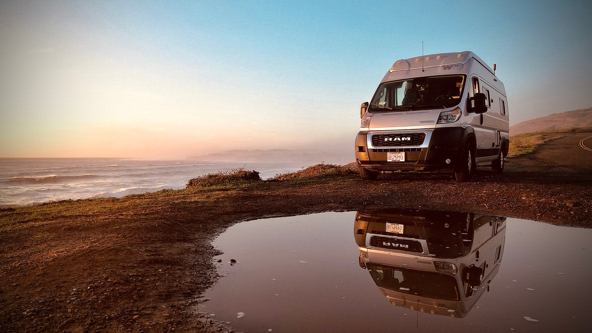 Coastal rv rental adventure! Silver camper van reflected in a puddle overlooking the ocean at sunset.