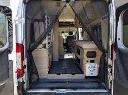 Inside an **rv rental** camper van, looking from the open rear doors through a mesh screen. Shows compact kitchen, storage, and clear aisle for mobile living.