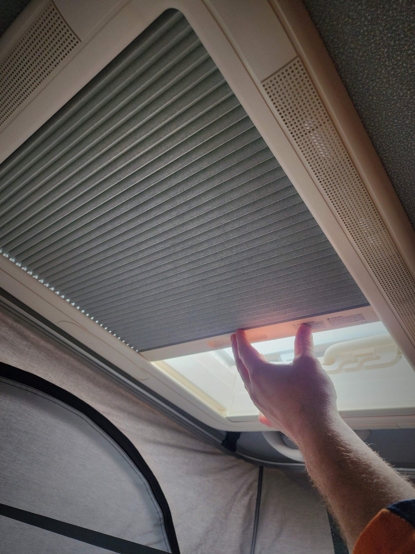 Hand adjusting a gray pleated skylight shade inside an RV rental. Light trim and ventilation grilles are visible.